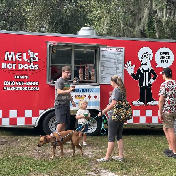 A family and their dog order food from the bright red Mel's Hot Dogs food truck.
