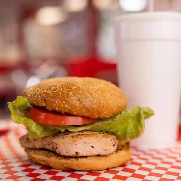 Grilled chicken sandwich with lettuce and tomato on a bun, next to a white cup.