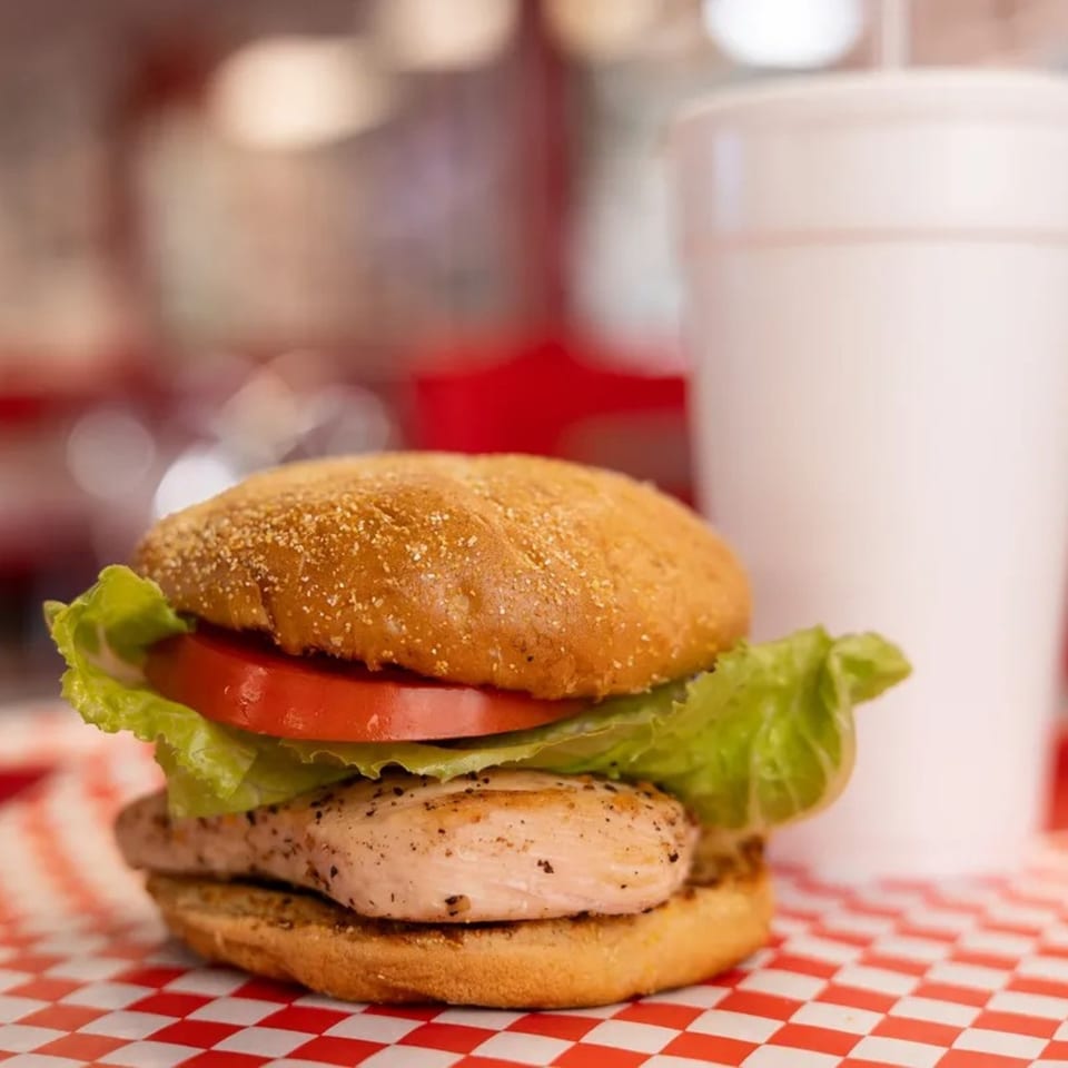 Grilled chicken sandwich with lettuce and tomato on a bun, next to a white cup.