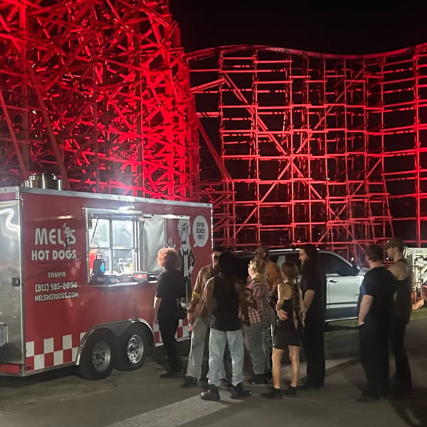 Mel's Hot Dogs food truck at night, with people gathered and a red-lit roller coaster in the background.