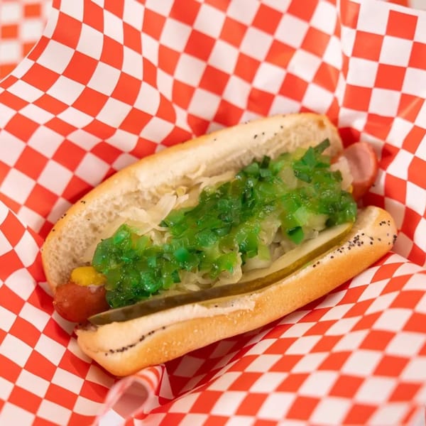 Mel's Hot Dog with green relish, mustard, and pickles in a red and white checkered paper.