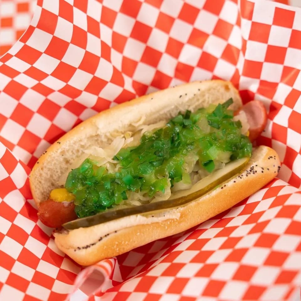 Mel's Hot Dog with green relish, mustard, and pickles in a red and white checkered paper.