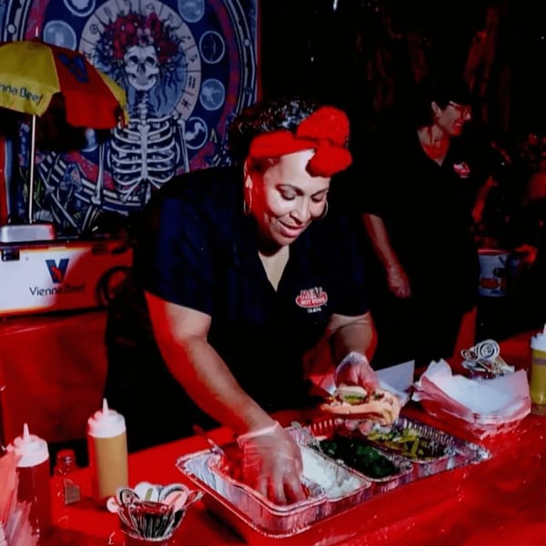 A Mel's Hot Dogs employee in a red bow prepares a hot dog at a vibrant red counter.