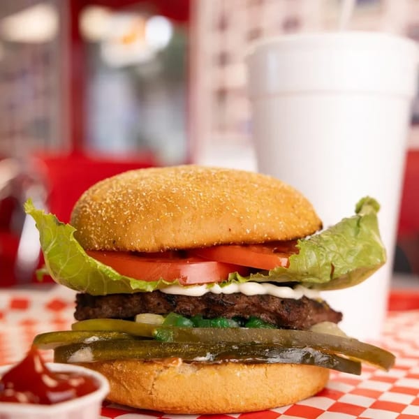 A Mel's Hot Dogs burger with lettuce, tomato, pickles, and relish on a checkered paper.