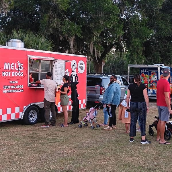 People line up at a red Mel's Hot Dogs food truck on a grassy field under trees.